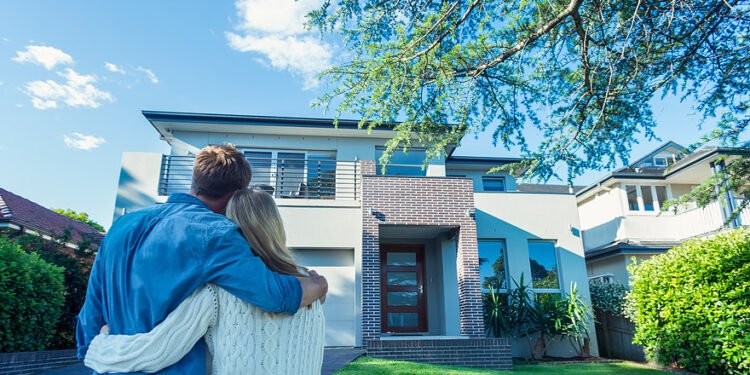 Couple standing in front of their new home. They are both wearing casual clothes and embracing. Rear view from behind them. The house is contemporary with a brick facade, driveway, balcony and a green lawn. The front door is also visible. Copy space